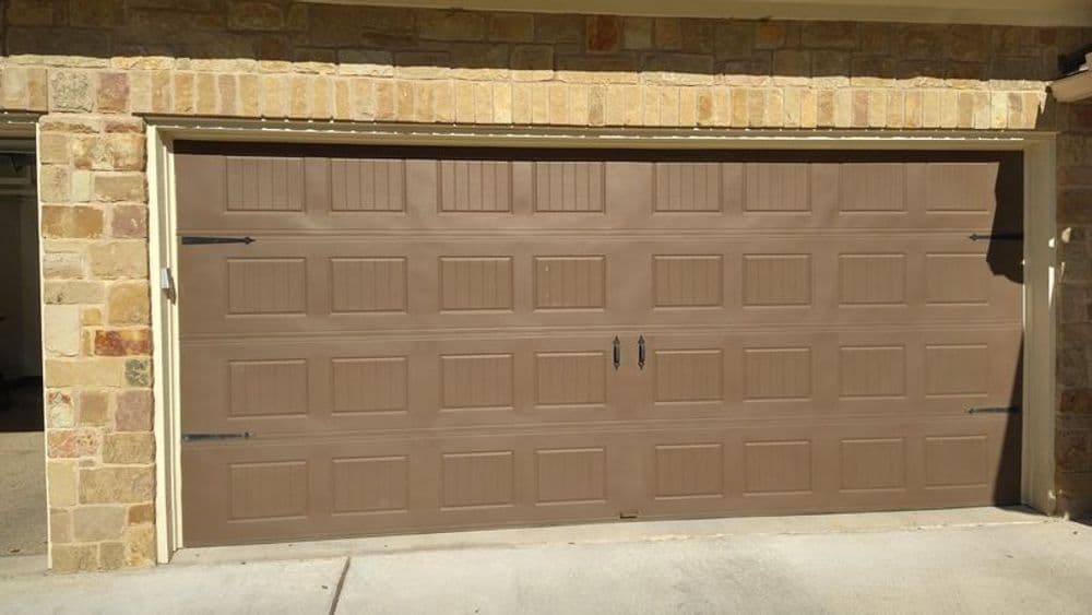 Brown insulated garage door with panel design and black handles on a stone wall.