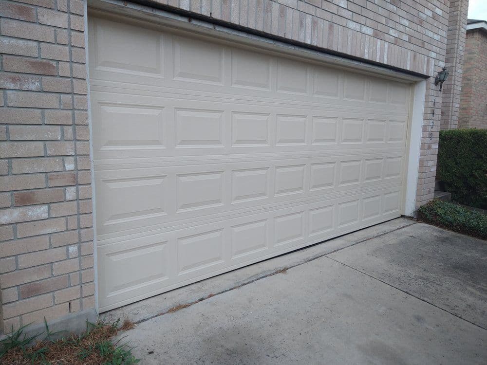 beige garage door on a brick house with a concrete driveway and green shrubbery nearby