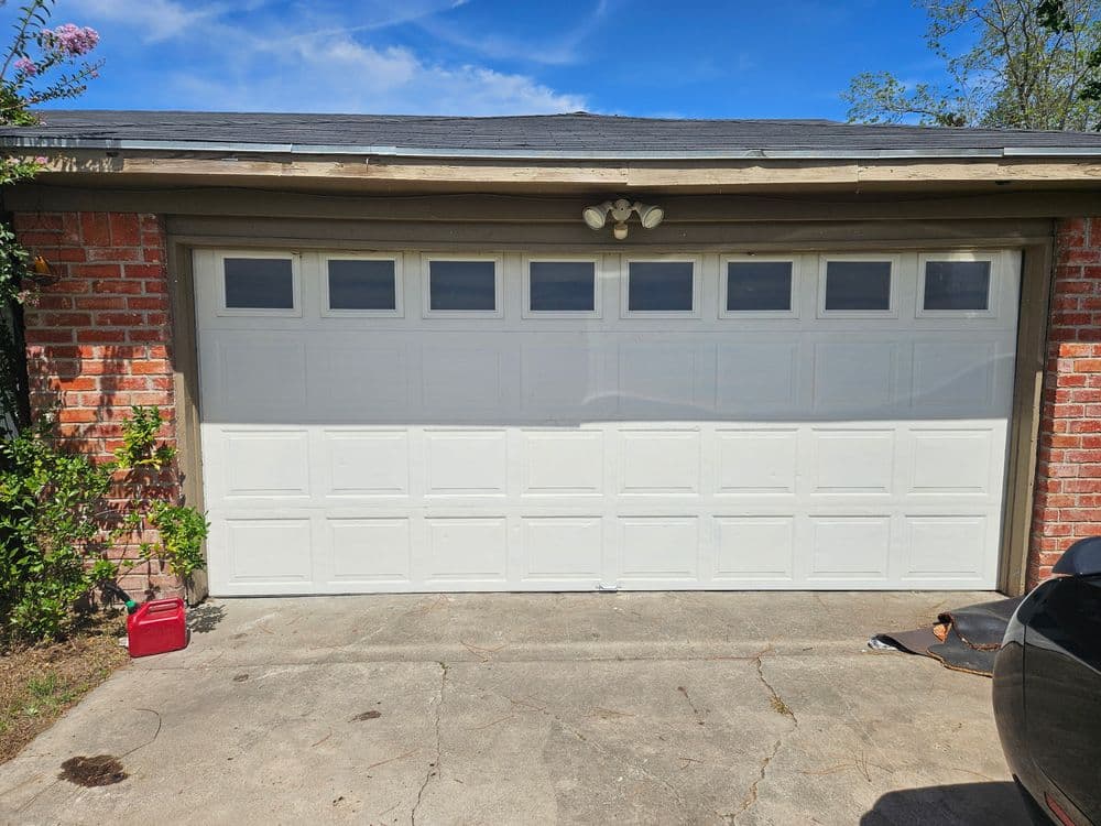 White garage door with windows on a brick house, surrounded by greenery and a concrete driveway.