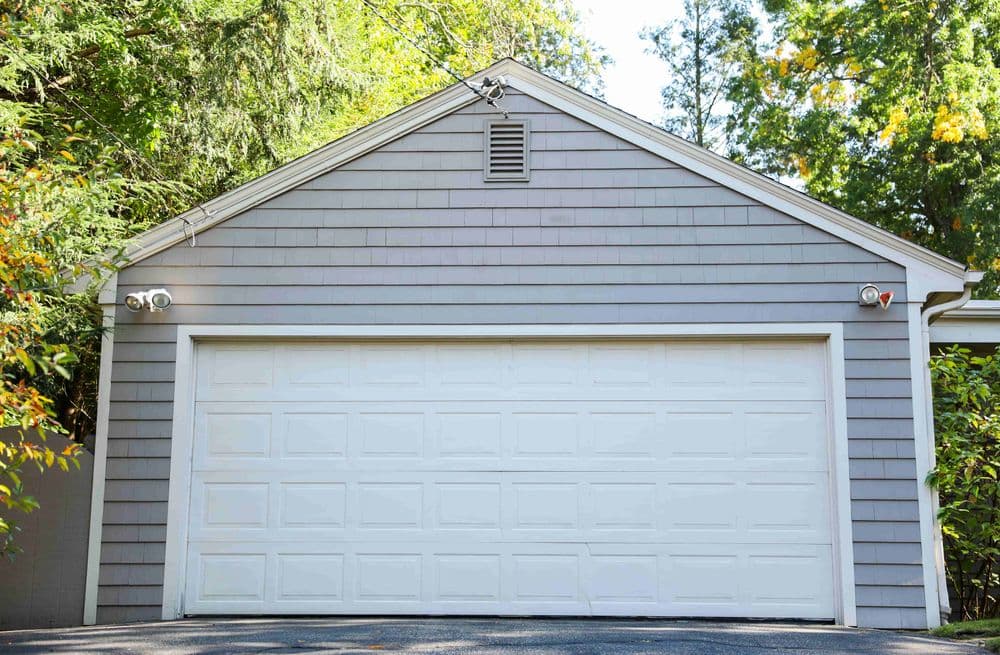 Gray garage door with decorative siding, surrounded by greenery and trees.