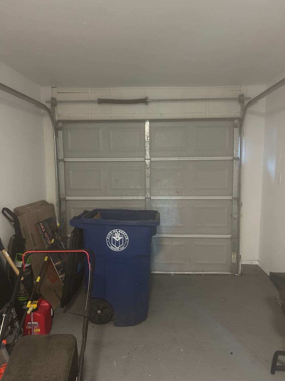Empty garage with a blue recycling bin, tools, and a lawn chair in the corner.