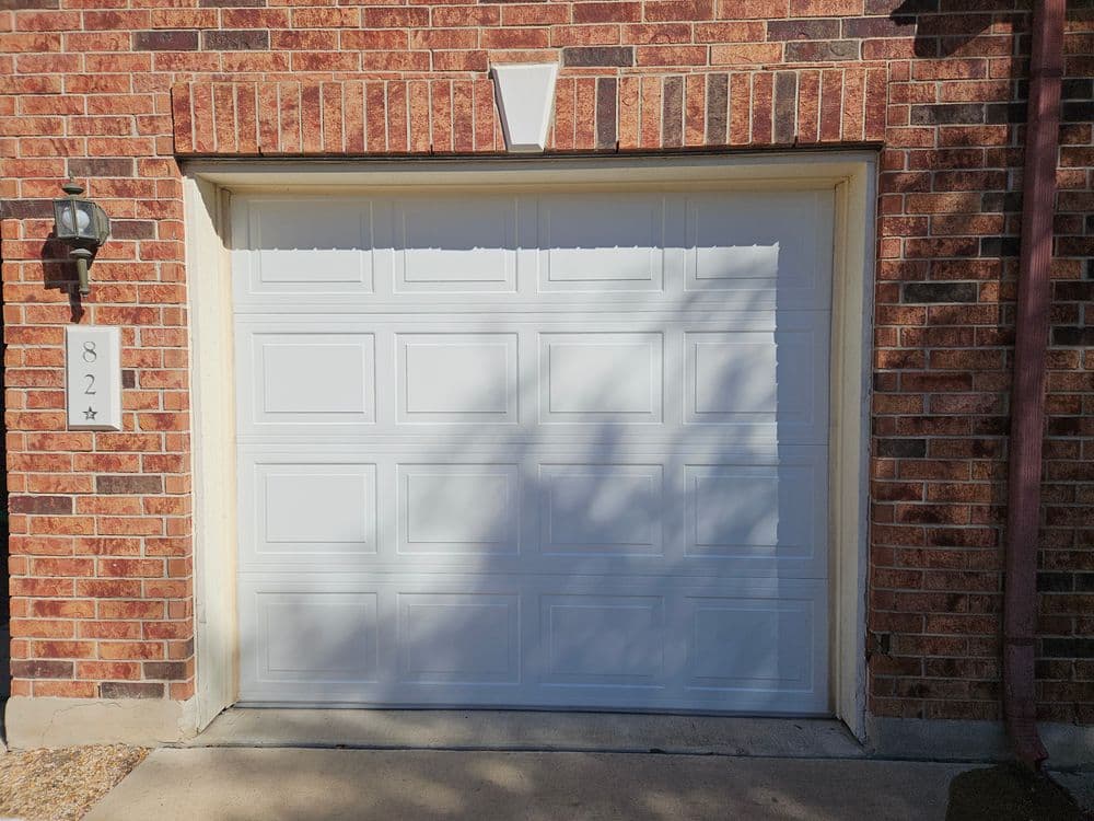 White garage door with brick wall, house number 82, and wall-mounted lamp.
